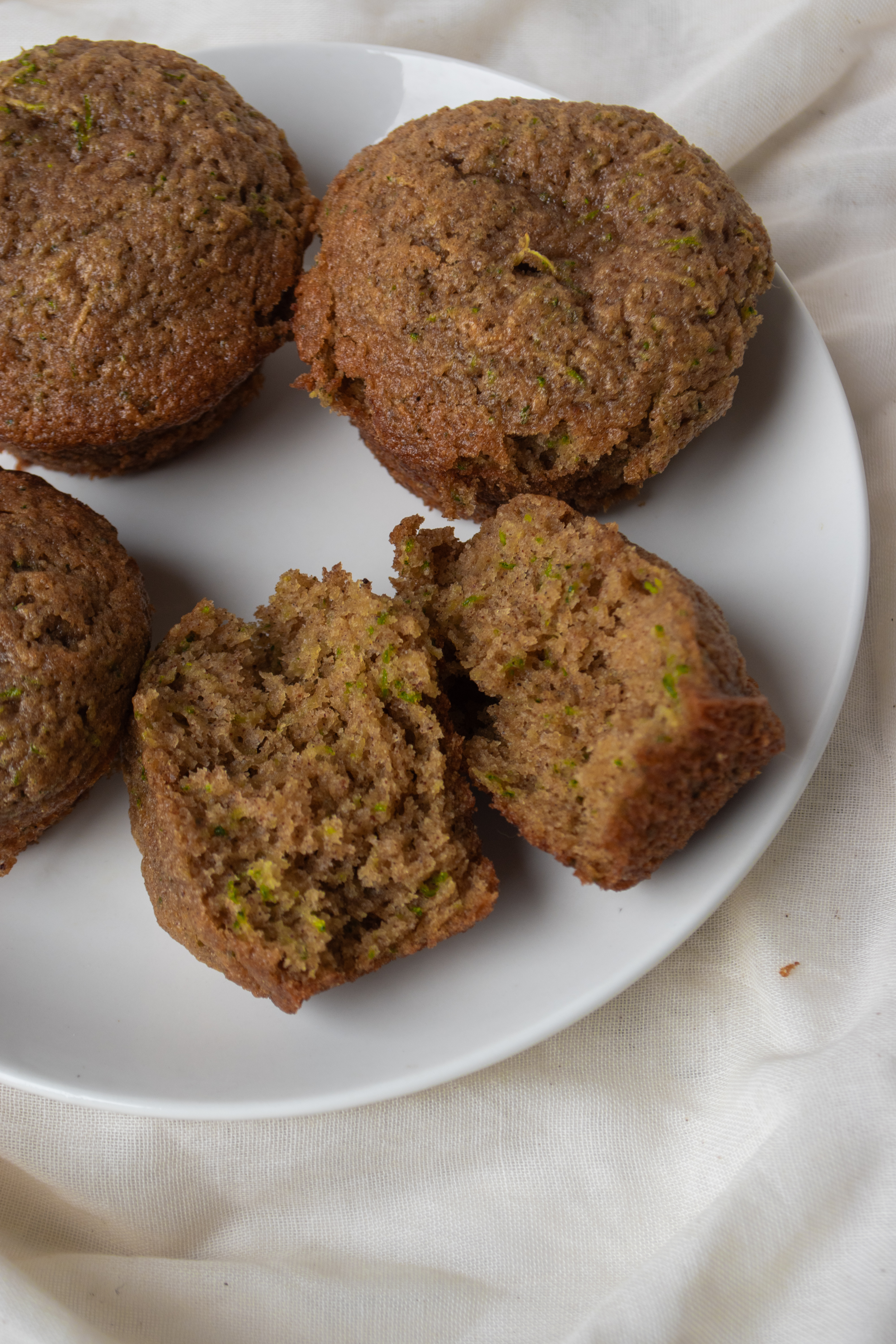 A plate with freshly baked muffins, displaying a few whole muffins and one cut open to show its moist texture and green flecks.